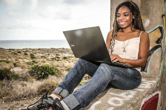 young woman sitting with her back against a tree working on her laptop