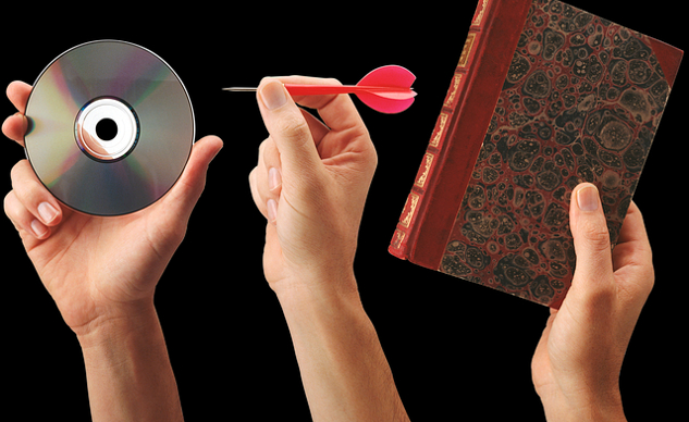 a hand holding a book, another hand holding a dart aimed at a CD with a hole in the center